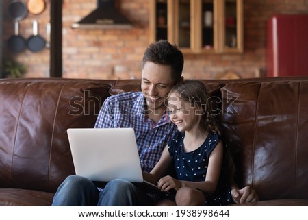 Laughing father and little daughter using laptop at home together, sitting on cozy couch in living room, happy dad and adorable child girl looking at screen, shopping, chatting online, watching video