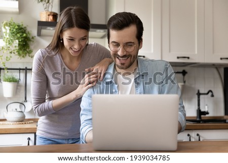 Happy young loving family couple looking at laptop screen, reading e-mail with good news. Smiling handsome man showing funny video on computer to pretty joyful wife, sitting at table in kitchen.