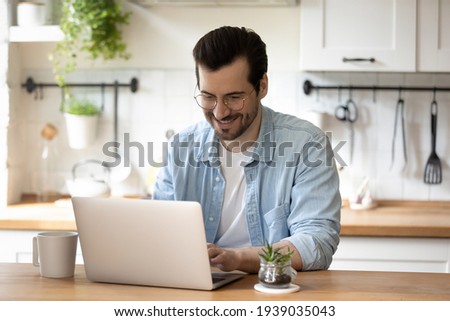Smiling young guy in glasses enjoying online communication, typing message in social networks on computer. Happy millennial male client involved in using software application, people and technology. Royalty-Free Stock Photo #1939035043