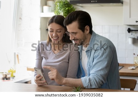 Pretty young woman holding smartphone in hands, showing funny photo editing application or online game to smiling handsome man, using electronic device together at free leisure time at home. Royalty-Free Stock Photo #1939035082