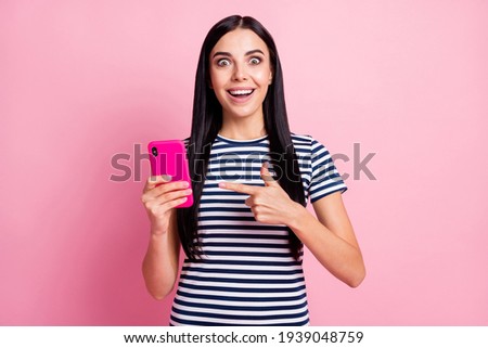 Photo portrait of amazed woman pointing finger at holding phone in one hand isolated on pastel pink colored background