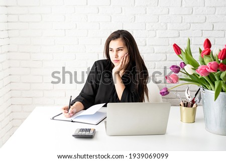 A young business woman florist writing in notebook, using laptop in the office, bucket of tulips on the desk. Small business.