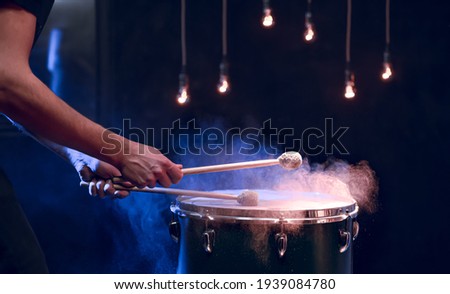 The percussionist plays with sticks on the floor tom on under studio lighting.. Concert and performance concept. Royalty-Free Stock Photo #1939084780
