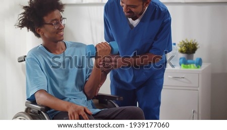 Indian physiotherapist helping disabled young man in wheelchair to work out with dumbbells. Handicapped afro-american patient doing rehabilitation exercises with nurse assistance Royalty-Free Stock Photo #1939177060
