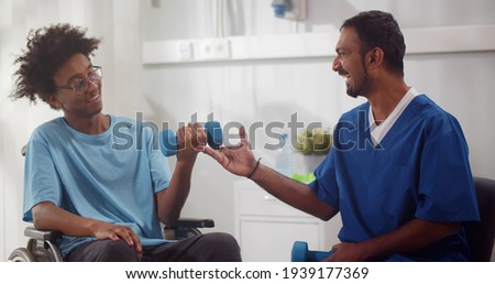 Healthcare worker and young afro patient in wheelchair during physiotherapy in hospital. Physiotherapist doctor helping to raise dumbbell to disabled afro man in wheelchair Royalty-Free Stock Photo #1939177369