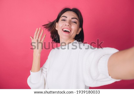 Beautiful woman in casual white sweater on pink red background do selfie on phone with huge smile excited hair flying isolated copy space