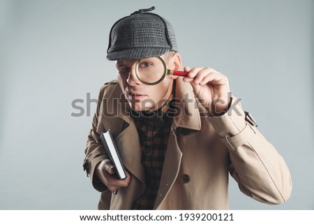 Male detective looking through magnifying glass on grey background Royalty-Free Stock Photo #1939200121