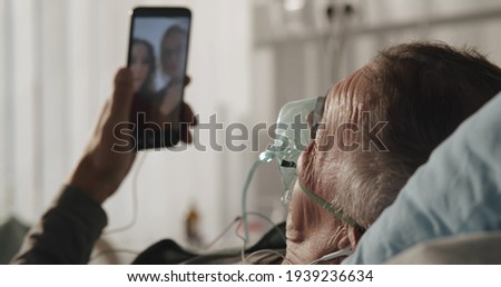 Patient is lying in bed wearing oxygen mask having video call with family in hospital ward. Sick aged male resting in hospital bed and having video conference on mobile phone Royalty-Free Stock Photo #1939236634