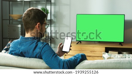 Caucasian man in glasses sitting on sofa in modern room in house watching TV with green screen and typing on smartphone with blank screen browsing online. Back view, leisure concept Royalty-Free Stock Photo #1939255126