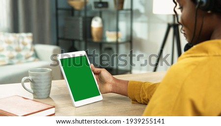 Close up of African American young pretty woman speaking in headset on video call on tablet device with chroma key while sitting at table in room in house. Video chat on gadget with green screen