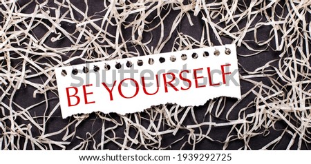 A white sheet of paper with the text BE YOURSELF lies on white shavings on a dark background.