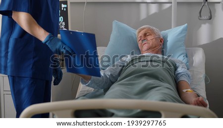 Sick senior man lying in hospital bed and signing informed consent. Medical worker in protective gloves holding clipboard while aged patient signing it lying in hospital ward