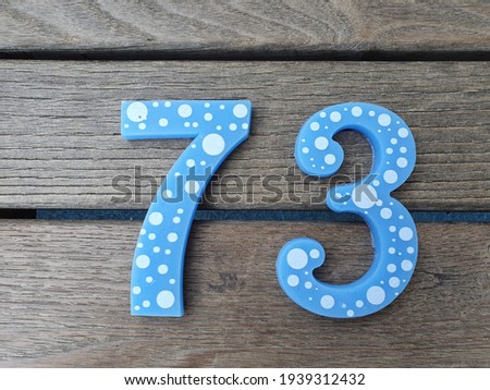 Blue Number 73 on brown wooden boards background. Light blue circles or bubbles or round shapes on the number seventy three. Cracks, slits, notch, scratches in wood planks.