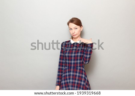 Studio portrait of tired exhausted young blond woman wearing checkered dress, raising and waving hand, puffing cheeks, trying to explain something, standing over gray background