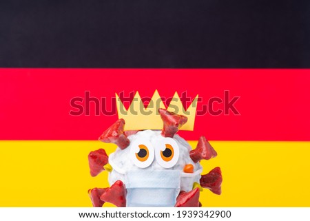 Coronavirus pandemic in Germany. Handmade model of Covid-19 cell with a crown, wearing medical mask in front of flag of Germany.
