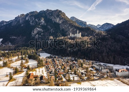 Aerial drone shot of Neuschwanstein Castle on snowy hill in winter sunlight in Germany with view of snowy village