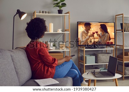 Woman enjoying good romcom serial in the evening. Single African American lady sitting alone at home, drinking coffee and watching romantic love story on soap opera entertainment TV channel, back view Royalty-Free Stock Photo #1939380427
