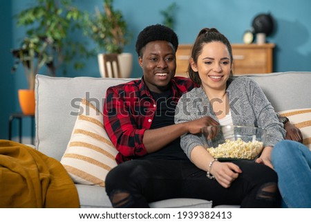 Cheerful Caucasian young couple laughing and talking while watching TV together on the couch athome with a bowl of popcorn.