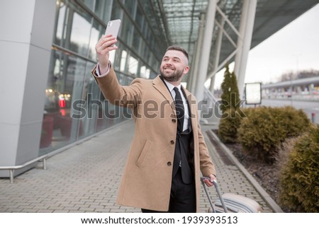 Mature male businessman, with beard, in black suit and coat, posing against airport background holding travel suitcase, and using phone