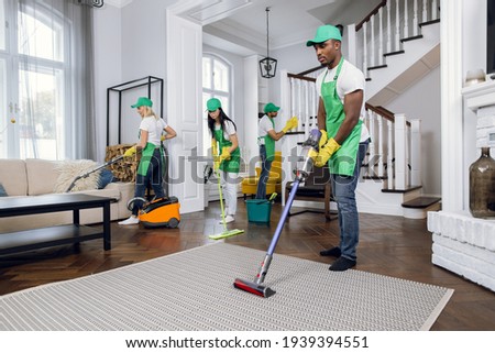 Cleaning team, group of four multiracial people in green aprons, hats and yellow gloves wiping, washing and vacuuming client house. Professional cleaning service. Royalty-Free Stock Photo #1939394551