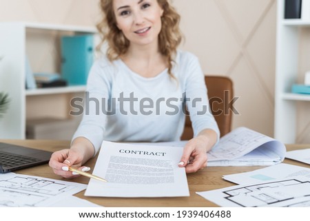 Realtor sitting in office behind desk, make offer to sign agreement for buy, rent or sale apartment. Woman holding document and pen in hands, showing paper on camera. Concept of building new house