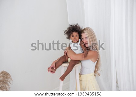 Portrait of young blond Caucasian mother and African American happy curly daughter on white background. A girl stands on a wooden chair, a woman hugs, kisses a baby. Adoption, mixed ethnic family.
