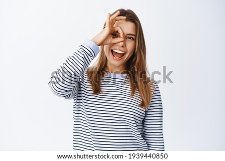 Excited beautiful woman laughing and showing okay sign on eye, checking out something cool, saying yes to good promo offer, standing over white background