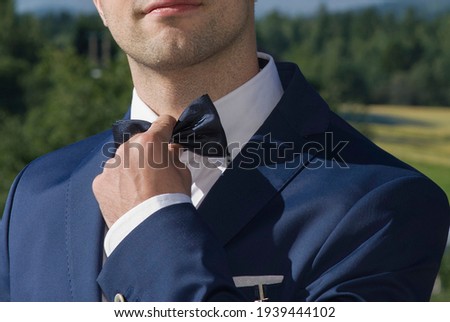 A smiling young man in a dark blue suit and white shirt, fixing a bow tie with his hand. Unrecognizable. No face. Blurred background with nature. Sunlight.