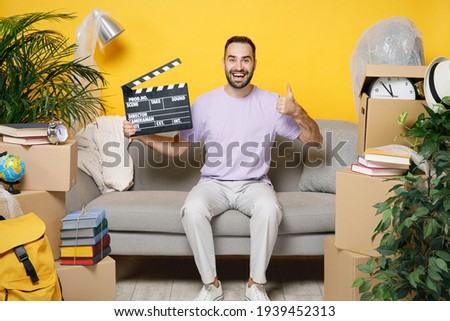 Smiling owner man showing thumb up hold film making clapperboard sit in living room on sofa at home household unpacking stuff rent flat isolated on yellow wall. Relocation moving in apartment concept Royalty-Free Stock Photo #1939452313