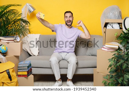 Excited owner man point thumb on himself doing selfie shot on mobile phone sit in living room on sofa at home unpacking stuff rent flat isolated on yellow wall. Relocation moving in apartment concept