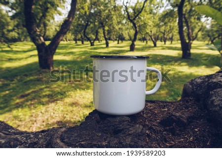 Enamel white mug on apple tree branch mockup. Stock countryside photo with white metal cup. Rustic scene, product mock up template. Lifestyle outdoors, trekking and camping design.