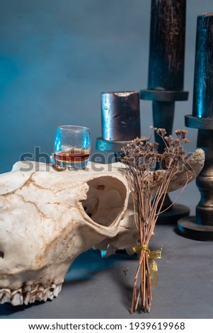 A glass of cognac stands on the skull of a bull next to black candles against a backdrop of blue smoke