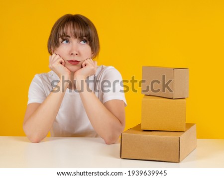 Woman thought about it. Girl in a pensive pose. Several boxes next to her. Girl dreams of her own business. Boxes as a symbol of commodity business. Portrait of a woman on a yellow background.