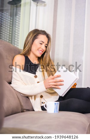 Lifestyle at home, young blonde Caucasian woman with a coffee smiling reading a book on the sofa in her living room