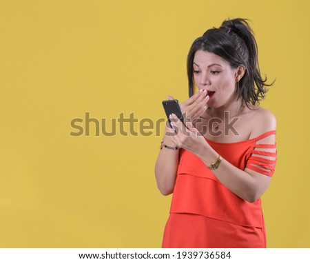 Surprised girl with smartphone in hand. Half-tail hairstyle. Orange blouse. Yellow background.