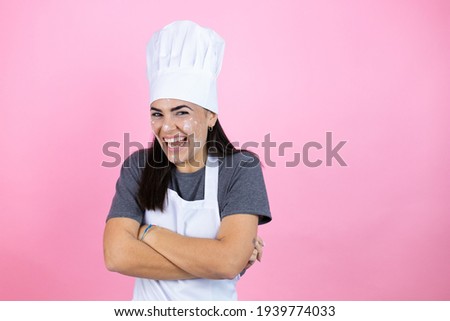 Young hispanic woman wearing baker uniform with flour on the face over pink background with a happy face standing and smiling with a confident smile showing teeth with arms crossed