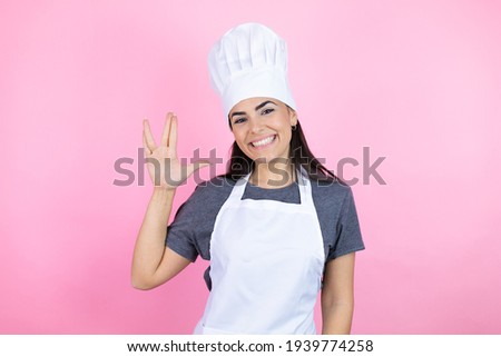 Young hispanic woman wearing baker uniform over pink background doing hand symbol