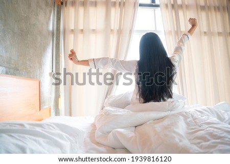 Happy Asian Young Girl Greets New Day with Warm Sunlight Flare, Sun shines on her from the big window.  Royalty-Free Stock Photo #1939816120