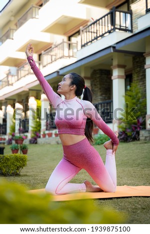 Sporty young healthy woman practicing yoga in garden, training on mat with plants and buildings on background.