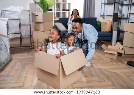 Young funny african father moving cardboard box with his little cute two daughters that sitting inside. Happy mother working on modern laptop on background. New flat for african family.