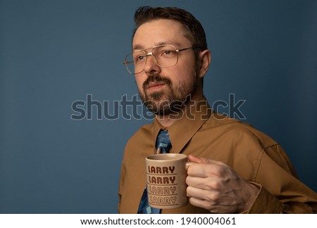 Studio portrait of a caucasian man dressed from the 1980s. He is wearing a buttoned up shirt and a vintage time and holding a coffee cup. The background is blue.