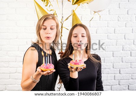 Birthday party. Two smiling young women or sisters in birthday hats celebrating birthday holding donuts with candles over white brick wall background