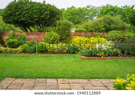 Backyard English cottage garden, colorful flowering plant and green grass lawn, brown pavement and orange brick wall, evergreen trees on background, in good care maintenance landscaping in park  Royalty-Free Stock Photo #1940112889