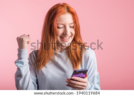 Redhead girl looks at the phone and shows the gesture of the winner in the studio on a pink background