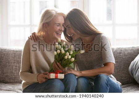 Loving young adult female child congratulate excited elderly mother with birthday anniversary at home. Smiling caring grownup millennial daughter present gift flowers to old mom on women s day. Royalty-Free Stock Photo #1940140240