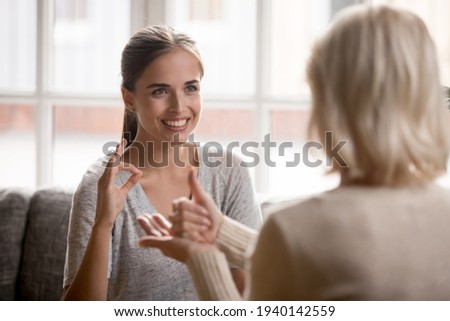 Smiling millennial grownup daughter use sign language make gesture talk communicate with older mom at home. Happy adult disabled impaired young woman have session with speech therapist or coach.