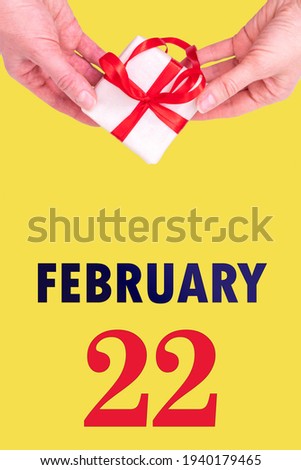 February 22nd. Festive Vertical Calendar With Hands Holding White Gift Box With Red Ribbon And Calendar Date 22 February On Illuminating Yellow Background. Winter month, day of the year concept.