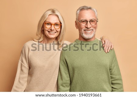 Smiling mature couple embrace look gladfully at camera pose for family portrait happy children came to visit them wear transparent glasses casual jumpers isolated over brown background. Age concept