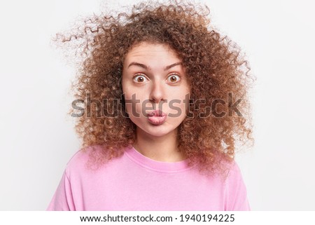 Portrait of surprised beautiful young European woman keeps lips folded looks amazed at camera has curly bushy hair dressed casually isolated over white background. Human face expressions concept