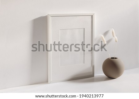 Modern summer stationery still life scene. Beige ball shaped vase with dry lagurus grass in sunlight with long shadows. Empty black vertical picture frame mockup. White table and wall background. 